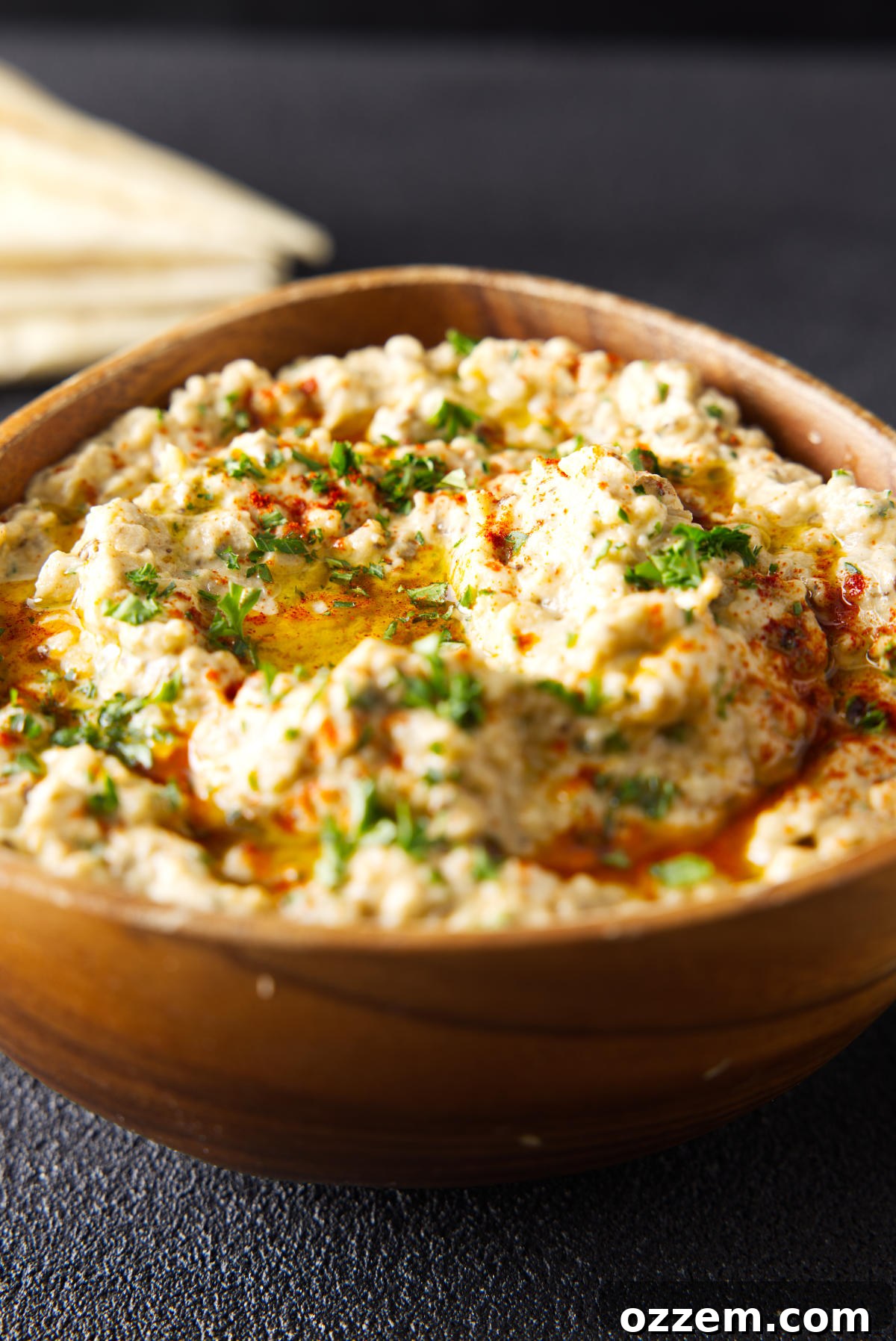 Wooden bowl filled to the brim with baba ganoush and topped with a drizzle of olive oil and chopped parsley, ready to be served.