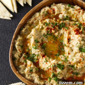Close up of smoked baba ganoush in wooden bowl topped with paprika, chopped parsley, and an olive oil drizzle.