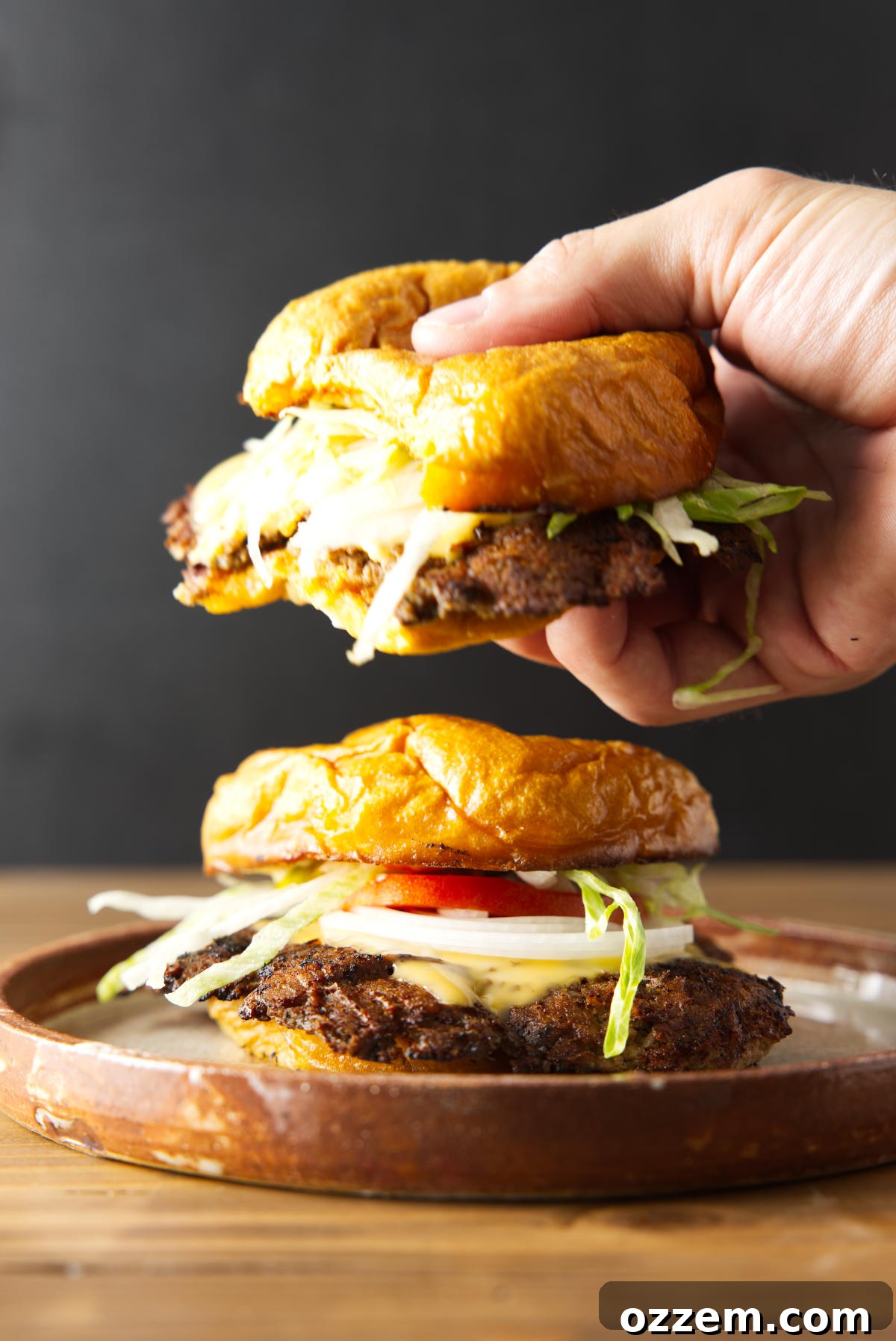 Blackstone Smash Burgers with Perfectly Crispy Edges 3 Holding a smash burger hovering over another plated smash burger loaded with toppings.
