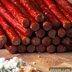 Close up of blue cheese venison snack sticks on butcher block with blue cheese crumbles and bacon in foreground.