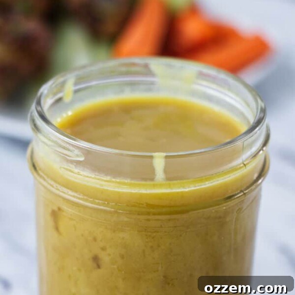 brownish yellow honey mustard sauce in mason jar with chicken wings and carrots and celery on a white plate in the background