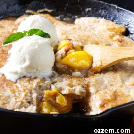 A spoon scooping peach cobbler from a cast iron skillet with vanilla ice cream.